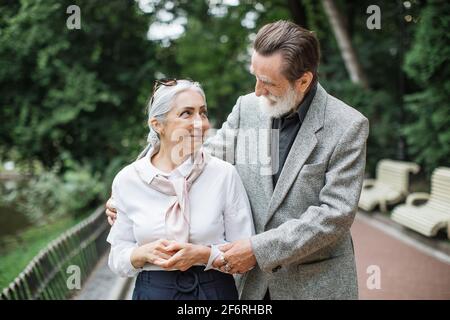 Glücklicher älterer Mann und charmante Dame, die im Freien lächelt und umarmt. Reizende, reife Familie, die in stilvoller Kleidung gekleidet ist und gemeinsam im grünen Park spazieren geht. Stockfoto