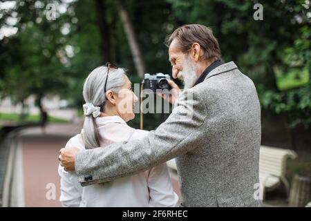 Rückansicht eines älteren Paares, das in Umarmungen im grünen Park steht und mit einer altmodischen Kamera fotografiert. Konzept von Familie, Ruhestand und Beziehung. Stockfoto