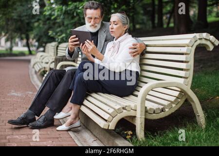 Glückliches reifes Paar mit digitalem Tablet, während es in Umarmung auf einer Holzbank sitzt. Entspannung im grünen Park. Konzept der modernen Technologie. Stockfoto