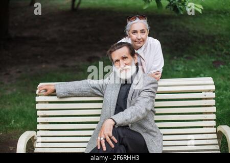 Attraktive kaukasische Rentner, die in eleganten Kleidern gekleidet sind und sich während der Freizeit im Park entspannen. Glückliche, reife Familie, die Zeit miteinander verbringt. Stockfoto