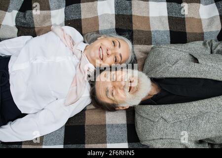 Draufsicht auf das ältere kaukasische Paar, das Kopf an Kopf auf einer karierten Decke lag und vor der Kamera lächelte. Frohe Ruhestand. Liebe seit Ewigkeiten. Stockfoto