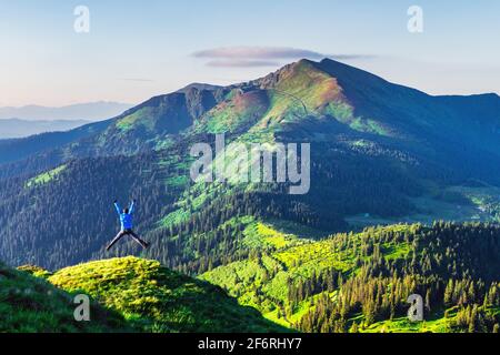 Ein Touristensprung am Rande eines Berges, der mit einem üppigen Gras bedeckt ist. Lila Sonnenuntergang Himmel und hohe Berge Gipfel auf dem Hintergrund. Landschaftsfotografie Stockfoto