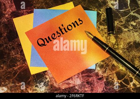 Wortschreiben TEXTFRAGEN auf einem Stapel mehrfarbiger Memo-Aufkleber, die mit einem schwarzen Füllfederhalter auf einem Marmortisch liegen. Stockfoto