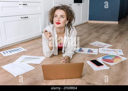 Glücklich nachdenkliche Freiberuflerin oder Studentin nutzt Laptop, arbeitet oder studiert zu Hause und lächelt. Erfolgreiche Geschäftsfrau liegt auf dem Boden Stockfoto