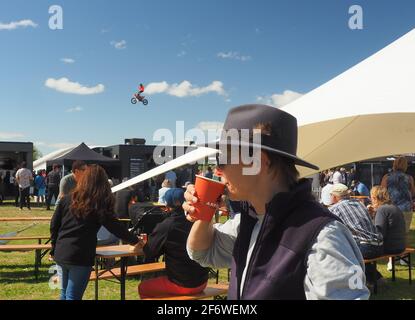 Motocross-Fahrer, der einen Sprung am 2021 Field Day in Feilding NZ durchführt. Eine Frau trinkt Kaffee zum Mitnehmen mit dem Reiter im air.behind. Stockfoto