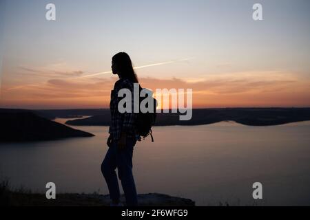 Silhouette einer jungen Frau in legerer Kleidung mit Rucksack auf der Schulter, der auf dem Berggipfel steht. Hintergrund des wunderschönen Sonnenuntergangs über der Bucht von Bakota. Stockfoto
