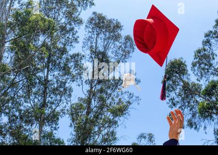 Hand eines Studenten, der seinen Abschlusshut und seine Maske über einen blauen Himmel auf dem Campus in die Luft wirft. Konzept von Graduierung und sozialer Distanzierung. Stockfoto