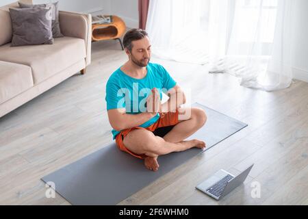 Mann mittleren Alters meditiert vor dem Laptop-Monitor. Yoga-Training zu Hause. Stockfoto