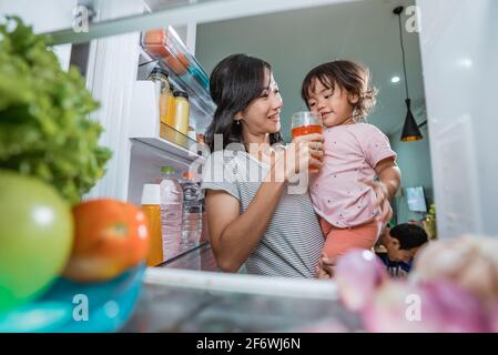 Mutter trägt ihre Tochter, während sie ein Glas Saft nimmt Im Kühlschrank Stockfoto