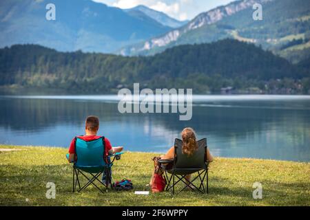 Pärchen sitzen entspannt auf Campingstühlen am Ufer des Hallstätter Sees oder des Hallstätter Sees in Gosaumühle, Österreich Stockfoto