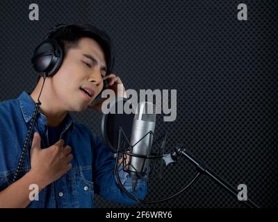 Junger asiatischer Mann mit Kopfhörern, der vor einer schwarzen Schallschutzwand singt. Musiker, der Musik im professionellen Aufnahmestudio produziert. Stockfoto