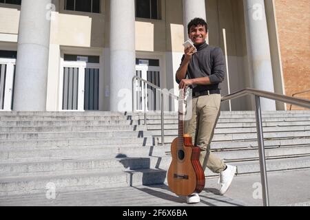 Ein junger lateinischer Mann lächelt, während er eine Tonnote auf seinem Smartphone aufzeichnet. Er hat eine Gitarre und steht auf einer Treppe. Stockfoto