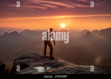 Behinderter Tourist mit Krücken. Rückansicht des verletzten Wanderers mit Unterarmstangen, die vor dem Hintergrund des Sonnenuntergangs am Berg laufen Stockfoto