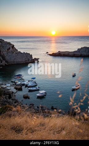 Saint Paul's Bay Strand mit Booten in Lindos, Rhodos, Griechenland bei Sonnenaufgang. Aufgehende Sonne zwischen zwei Klippen Stockfoto