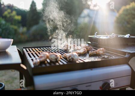 Nahaufnahme eines bbq-Grills mit Fleisch auf ihnen kochen, Sonneneinstrahlung im Hintergrund fängt den Rauch auf Stockfoto