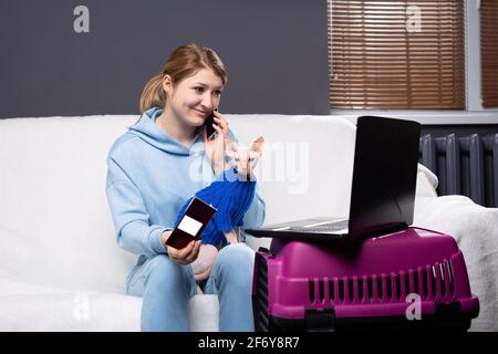 Schöne lächelnde junge Frau, die mit ihrem Katzentier spielt, während sie einen Laptop-Computer benutzt und auf dem Smartphone spricht. Mädchen bereiten Online-Ticket Pässe für Stockfoto