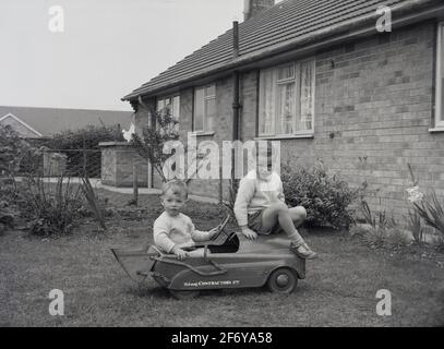 1956, historisch, außerhalb eines bung]alow in einem Garten, zwei kleine Jungen auf einem Tri-ang-Metall-Ride-On-Spielzeugauto, einer auf dem Fahrersitz, der andere auf der Motorhaube sitzen. An der Rückseite des Fahrzeugs befindet sich ein Metalleimer oder Bagger und auf der Seite die Aufschrift „Tri-ang CONTRACTORS Ltd“. Tri-Ang war der Markenname des Spielzeugunternehmens Lines Brothers, das nach dem 1. Weltkrieg von drei Brüdern gegründet wurde und zu einer Zeit als das größte Spielzeugunternehmen der Welt gilt. Obwohl ihr Vater ein Spielzeuggeschäft hatte, Holzspielzeug auf Tierbasis, sahen die Lines Brothers, dass Kinder Metallfahrzeuge mit Rädern mögen. Stockfoto