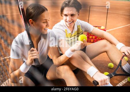 Fröhliches Paar, das auf dem Tennisplatz mit Schlägern und Ball in den Händen sitzt. Mädchen zeigt gelben Ball zu ihrem Tennis-Partner Stockfoto