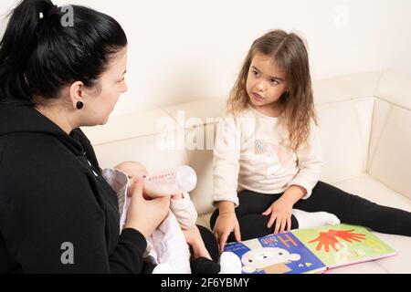 Vierjähriges Mädchen, das sich ein Bilderbuch ansieht und mit der Mutter spricht, die eine 2 Monate alte Schwester füttert Stockfoto