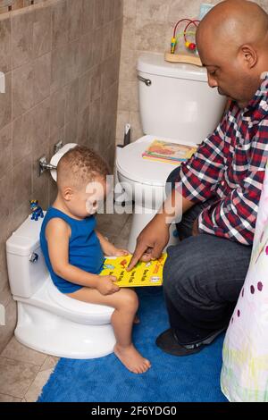 Toilettentraining: 2-jähriger Junge sitzt auf einem Töpfchen im Badezimmer und schaut sich mit dem Vater Bücher an Stockfoto