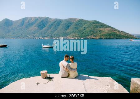 Ein Mann und eine Frau sitzen und umarmen am Rande eines Piers in der Nähe von Perast in der Bucht von Kotor, Rückansicht Stockfoto