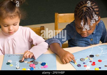 Bildung Vorschule 3-4-Jährige zwei Mädchen sitzen nebeneinander und machen Kunstaktivitäten kleben Knöpfe auf Baupapier, eines mit der rechten Hand und t Stockfoto