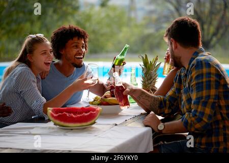 Zwei Paare, die am Tisch am Pool sitzen, reden, lächeln, lachen, sich unterhalten, Bier und Weißwein trinken Stockfoto