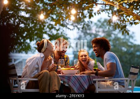 Gruppe multiethnischer Freunde, die lachen, sich versammeln, reden, am Tisch im Freien sitzen, sich unterhalten, Stockfoto