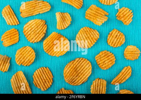 Kartoffelchips, gerillte Goldchips, knusprige Snacks aus Wellpappe auf türkisfarbenem Hintergrund, farbiges Muster. Ungesunde Fast Food. Textur mit gesalzenen Scheiben. Hig Stockfoto