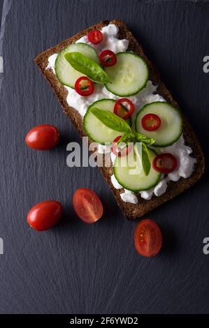 Frisches Frühlingsbrot auf einem glutenfreien Sonnenblumenbrot in Scheiben. Rotes und grünes Gemüse auf schwarzem Schieferstein. Gurke, Chili und Quark Garni Stockfoto