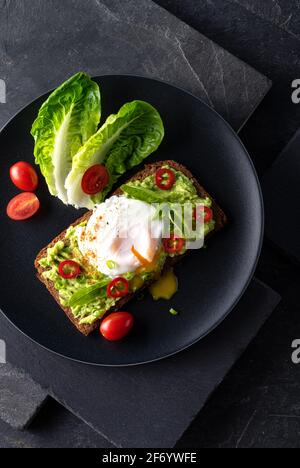 Frisches Frühlingsbrot auf einem glutenfreien Sonnenblumenbrot in Scheiben. Rotes und grünes Gemüse auf schwarzem Schieferstein. Pochiertes Ei, Avocado, Chili und Spring o Stockfoto