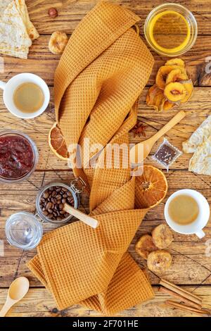 Zusammensetzung der Frühstückssnacks mit einem gefalteten Küchentuch, um das Pfannkuchen, Obstchips, Nüsse, Honig und zwei Tassen Kaffee herum gelegt werden. Gesunder Snack und Öko f Stockfoto