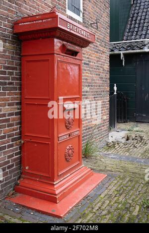 Historischer traditioneller roter Metallpostkasten, der an einer Ziegelmauer in Zaandijk, einem typisch kleinen Dorf in den Niederlanden, aufgestellt ist Stockfoto