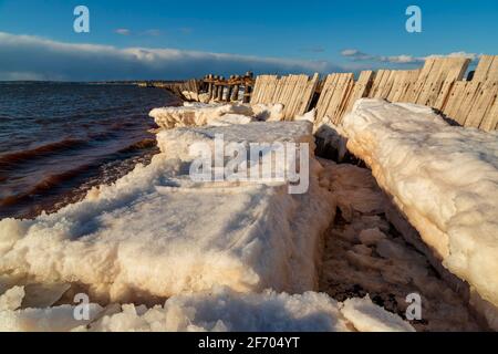 Alte und verfallene hölzerne Anlegestelle, die in gefrorenen Eisblättern gefangen ist Auf offenem Wasser Stockfoto