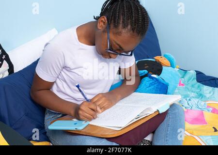 12-jähriges Mädchen mit Brille auf dem Bett sitzend Zu Hause Schularbeiten machen Stockfoto
