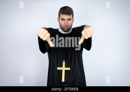 Junger hispanischer Mann in Priesteruniform, der auf weißem Hintergrund mit einem wütenden Gesicht steht, ein negatives Zeichen, das Abneigung mit Daumen nach unten zeigt Stockfoto