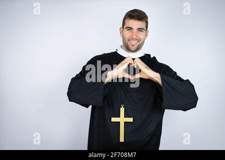 Junger hispanischer Mann in Priesteruniform, der auf weißem Hintergrund steht Lächelnd in der Liebe zeigt Herz-Symbol und Form mit den Händen Stockfoto