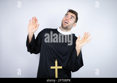 Junger hispanischer Mann in Priesteruniform, der auf weißem Hintergrund steht Erschrocken mit ihren Armen nach oben wie etwas, das von oben fällt Stockfoto