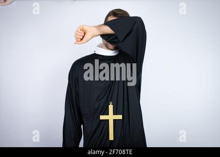 Junger hispanischer Mann in Priesteruniform, der auf weißem Hintergrund steht und die Augen mit einem lächelnden Arm bedeckt, fröhlich und lustig. Blindes Konzept. Stockfoto