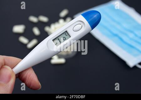 Hand hält weißes elektronisches Thermometer im Hintergrund mit Packungen von Tabletten und blauer medizinischer Gesichtsmaske. Selektiver Fokus. Stockfoto