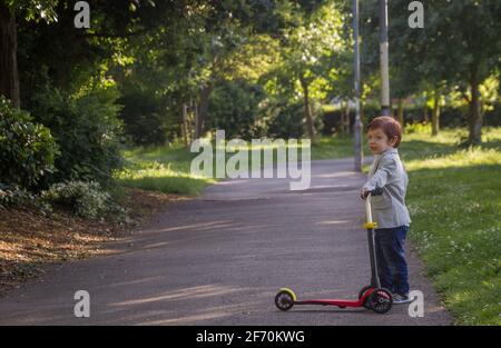 Ein süßer, rothaariger, blauäugiger Junge, der an einem sonnigen Tag einen roten Roller in einem Park hält und einen Blazer trägt Stockfoto