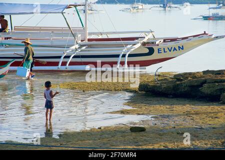 Ein philippinischer Fischer fährt zu seinem Boot, während er von einem Jungen beobachtet wird, der im flachen Wasser der Ebbe, Moalboal, Cebu Island, Philippinen, paddelt. Stockfoto