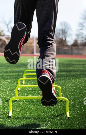 Rückansicht eines männlichen Athleten, der während des Trainings auf einem Bein über gelbe Mini-Hürden auf einem grünen Rasenfeld springt, aus einem niedrigen Winkel. Stockfoto