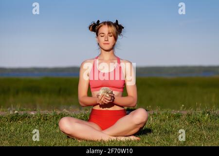 Im Sommer praktiziert die junge Frau mit ihrem Igel auf grünem Gras im Park Yoga-Meditation. Entspannen und Harmonie mit der Natur. Stockfoto