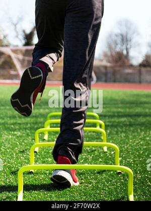 Low-Angle-Ansicht eines männlichen Athleten, der auf einem Bein über gelbe Mini-Hurldes auf einem grünen Rasenfeld von hinten springt. Stockfoto