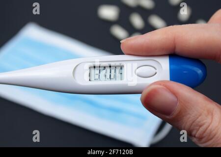 Hand hält weißes elektronisches Thermometer im Hintergrund mit Packungen von Tabletten und blauer medizinischer Gesichtsmaske. Stockfoto