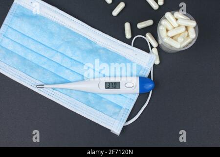 Weißes elektronisches Thermometer mit Packungen Tabletten und blauer medizinischer Gesichtsmaske auf schwarzem Hintergrund. Stockfoto