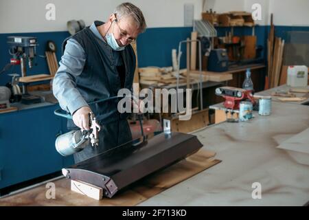 Ein pensionierter Mann in einer medizinischen Maske malt in einer Schreinerei mit einer Kompressorpistole Holzmöbel. Ein erfahrener Arbeiter malt das Teil sorgfältig. Stockfoto