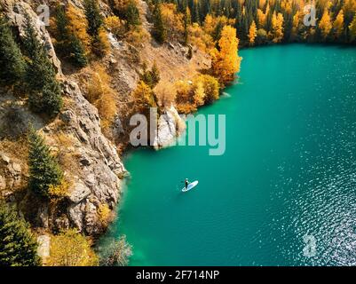 Luftdrohnenaufnahme von man, der im Herbst auf einem SUP-Brett am Bergsee in der Nähe eines gelben Waldes schwimmt. Abenteuer beim Stand Up Paddle Boarding. Stockfoto