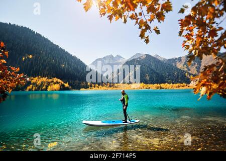 Mann schwimmt im Herbst auf einem SUP-Brett am Bergsee in der Nähe des gelben Waldes. Abenteuer beim Stand Up Paddle Boarding. Stockfoto
